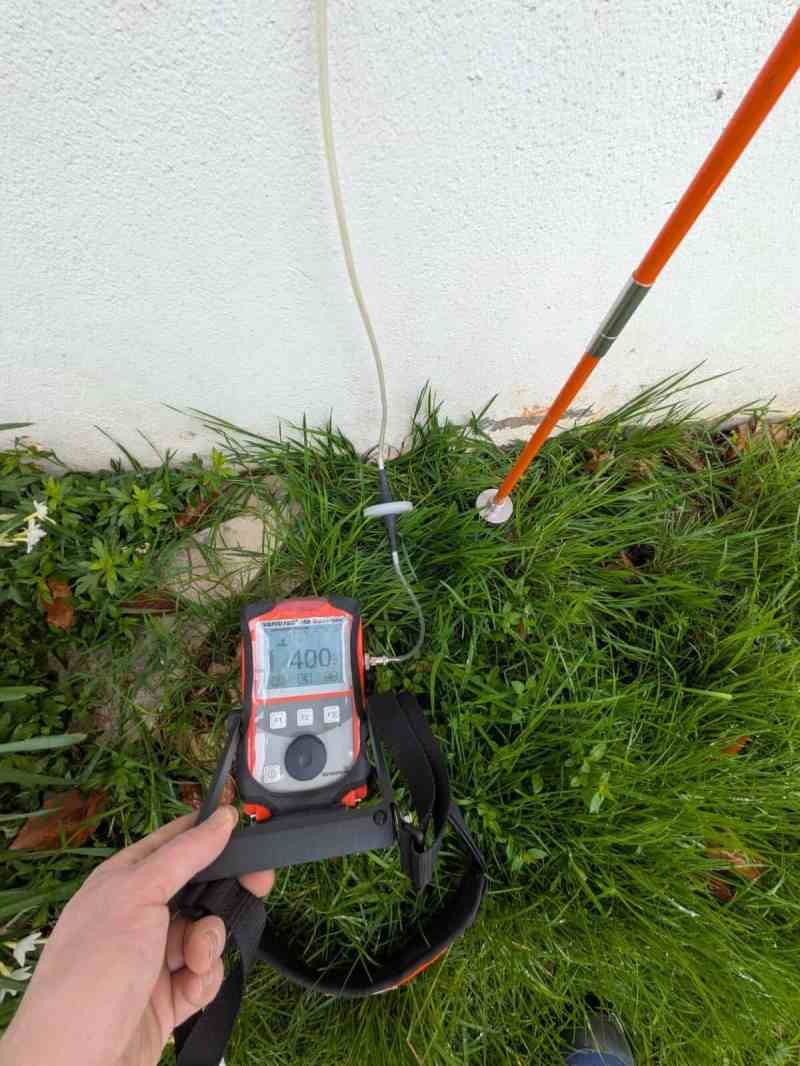 Détecteur de gaz traceur posé dans l’herbe au pied d’un mur, sonde à côté